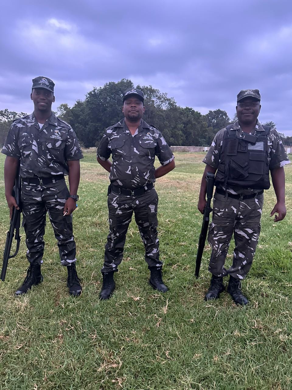 Three security officers in camouflage uniforms standing confidently on a grassy field, showcasing High Risk Protection's readiness and professionalism.
