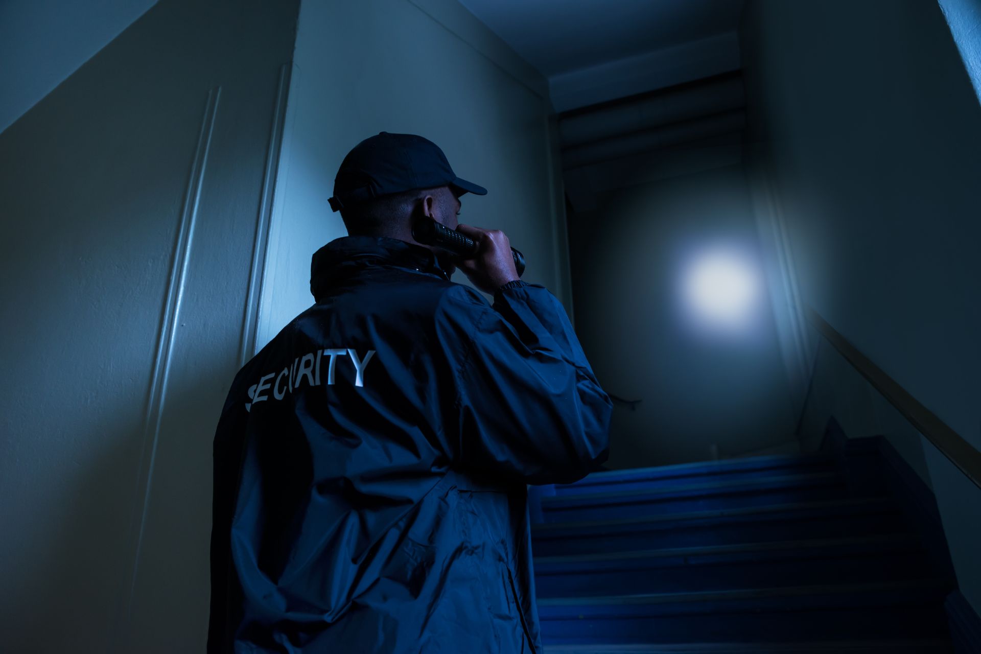 Security officer in a dark stairwell communicating on a radio, vigilant during a night patrol, representing professional vigilance by High Risk Protection.
