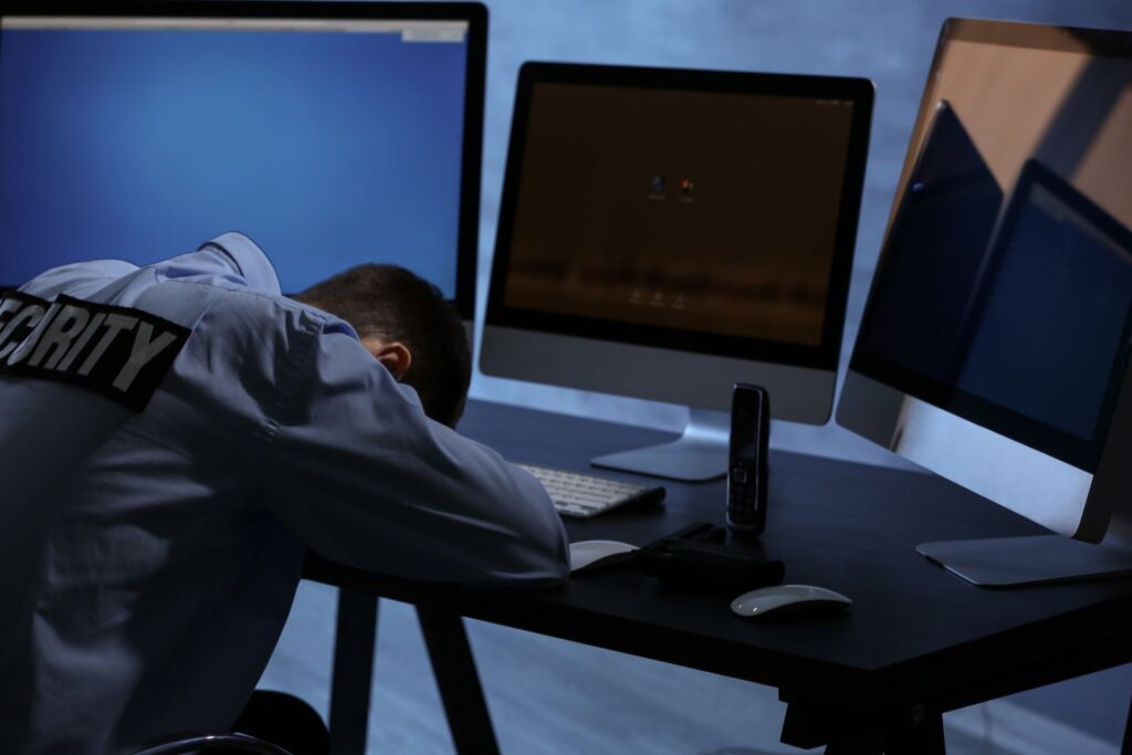 Exhausted security officer resting his head on a desk surrounded by multiple computer monitors during a night shift, highlighting the stressful nature of security work