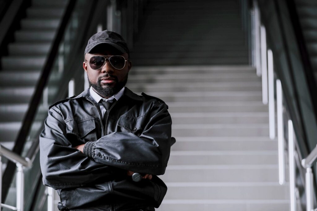 Professional security guard in Pretoria standing confidently with arms crossed, wearing a black jacket, sunglasses, and cap in an urban setting